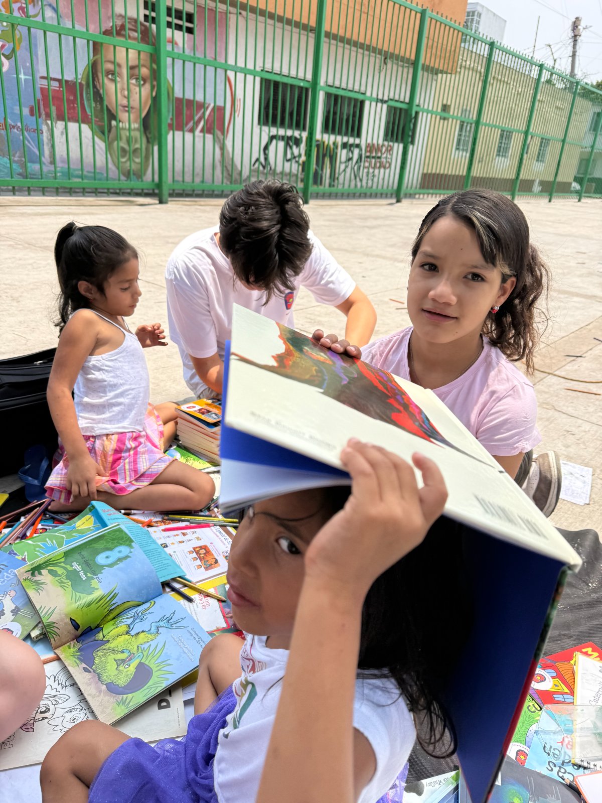 Children reading picture books together on a blanket in a courtyard, one holding an open book over her head