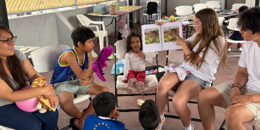Volunteers and children seated in a circle at a migrant shelter during library time, one volunteer holding an open picture book